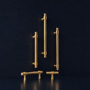 May include: A selection of gold-coloured cabinet pulls and handles in different sizes. The hardware has a cylindrical bar design with rounded ends and a T-shaped base. The pulls are set against a dark blue backdrop.