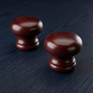 May include: Two round, dark reddish-brown wooden cabinet knobs with a smooth, polished finish. Each knob features a mushroom-shaped top and a tiered, cylindrical base, showcasing the natural wood grain texture throughout the surface of the hardware.
