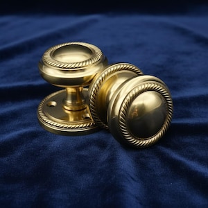 May include: Two brass door knobs with a rope design around the edges. One knob is attached to a round base, while the other is separate. The knobs are a shiny gold color and sit on a dark blue velvet surface.