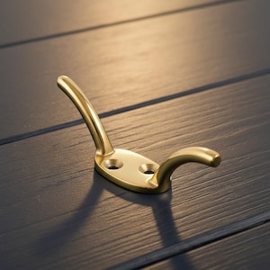 May include: A brass-coloured wall hook with a curved design. The hook has a flat base with two screw holes and a curved arm for hanging items. The hook is made of metal and has a shiny finish.