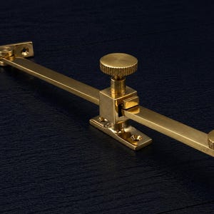 Può includere: Un fermavetro in ottone, con una base rettangolare e un braccio girevole con un pomello a ciascuna estremità. L'hardware è progettato per finestre o porte. La finitura è dorata e lucida.