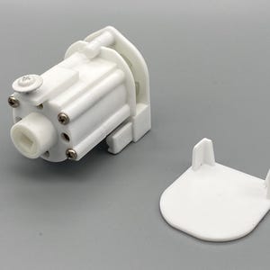 May include: A white plastic blind mechanism with a cylindrical body and a separate, matching cover. The mechanism has several small screws and a square opening. The cover has two vertical posts.