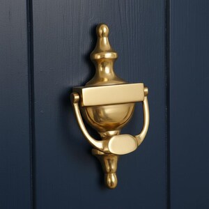 May include: A gold-coloured door knocker with an elegant urn design. The knocker has a rectangular plate, a curved handle, and a decorative top. It is mounted on a dark blue wooden door.