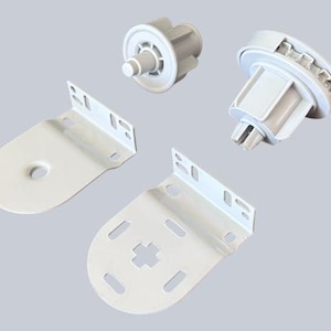 May include: An assortment of white roller blind components, including two brackets, and two cylindrical mechanisms. The brackets have a curved bottom with a hole and slots for mounting. The mechanisms have gears and a central pin.