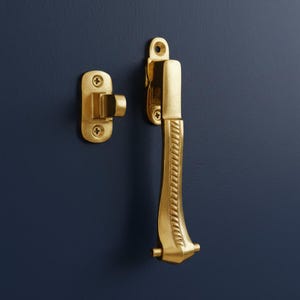May include: A set of two brass-colored window or cabinet hardware pieces. The hardware includes a handle with a decorative pattern and a matching latch, both mounted on a dark blue surface.