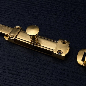 May include: A shiny, gold-colored sliding bolt latch with a round knob and a separate strike plate. The hardware is made of metal and is designed for securing doors or gates. The latch is shown against a dark blue background.