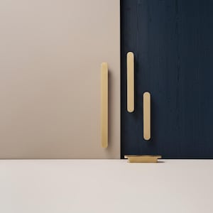 Peut inclure: Composition minimaliste avec plusieurs poignées de meubles dorées et un petit plateau rectangulaire. Les poignées sont longues et fines, avec des extrémités arrondies, disposées sur un fond de panneaux beiges et bleu marine. Le plateau est également doré.