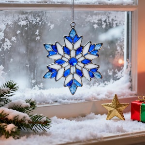May include: A stained glass snowflake ornament in shades of blue and white hangs in front of a snowy window. A gold star and a small wrapped gift sit on the windowsill. The ornament is a winter holiday decoration.