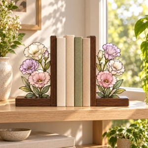 Puede incluir: Un par de sujetalibros de madera oscura en forma de L con paneles decorativos de vidrio tintado. Cada panel muestra un grupo de flores ilustradas en tonos blanco, lavanda y rosa con hojas verdes, sosteniendo cuatro libros de colores neutros.
