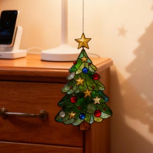 K&ouml;nnte beinhalten: Ein Weihnachtsbaumschmuck aus Glasmalerei. Der gr&uuml;ne Baum ist mit roten und blauen Kugeln und goldenen Sternen verziert. Ein goldener Stern ziert die Spitze des Baumes. Der Schmuck h&auml;ngt vor einem Nachttisch aus Holz.