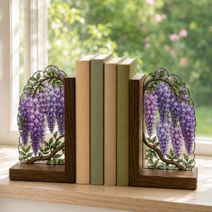 Könnte beinhalten: Ein Paar L-förmige Buchstützen aus Holz mit dekorativen Metalleinsätzen, die eine Illustration im Buntglas-Stil von violetten Glyzinienblüten und grünen Blättern zeigen und vier Bücher mit einfarbigen Buchrücken stützen.