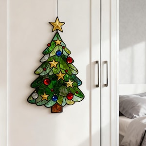 K&ouml;nnte beinhalten: Ein Weihnachtsbaumschmuck aus Glasmalerei. Der Baum ist gr&uuml;n mit goldenen Sternen, roten und blauen Kugeln und einem goldenen Stern an der Spitze. Der Baum h&auml;ngt vor einem wei&szlig;en Hintergrund.