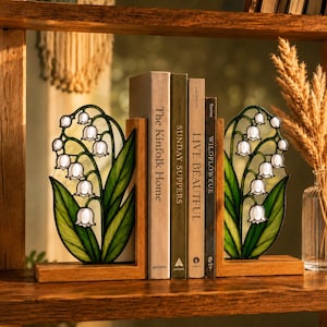 May include: A pair of wooden bookends featuring stained glass panels shaped like lily of the valley flowers with white bell-shaped blossoms and green leaves. The bookends support four books titled The Kinfolk Home, Sunday Suppers, Live Beautiful, and Wildflower.