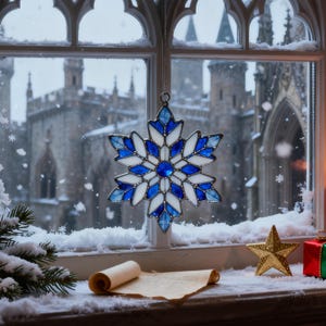May include: A blue and white stained glass snowflake decoration hangs in front of a window. The ornament is suspended by a chain. The window sill has snow, a gold star, a scroll, and a small gift box.