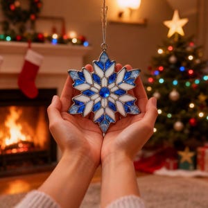 May include: A stained glass snowflake ornament with blue and white segments, held in cupped hands. The ornament has a silver hanging loop. The background includes a Christmas tree with lights and a fireplace with a fire burning.