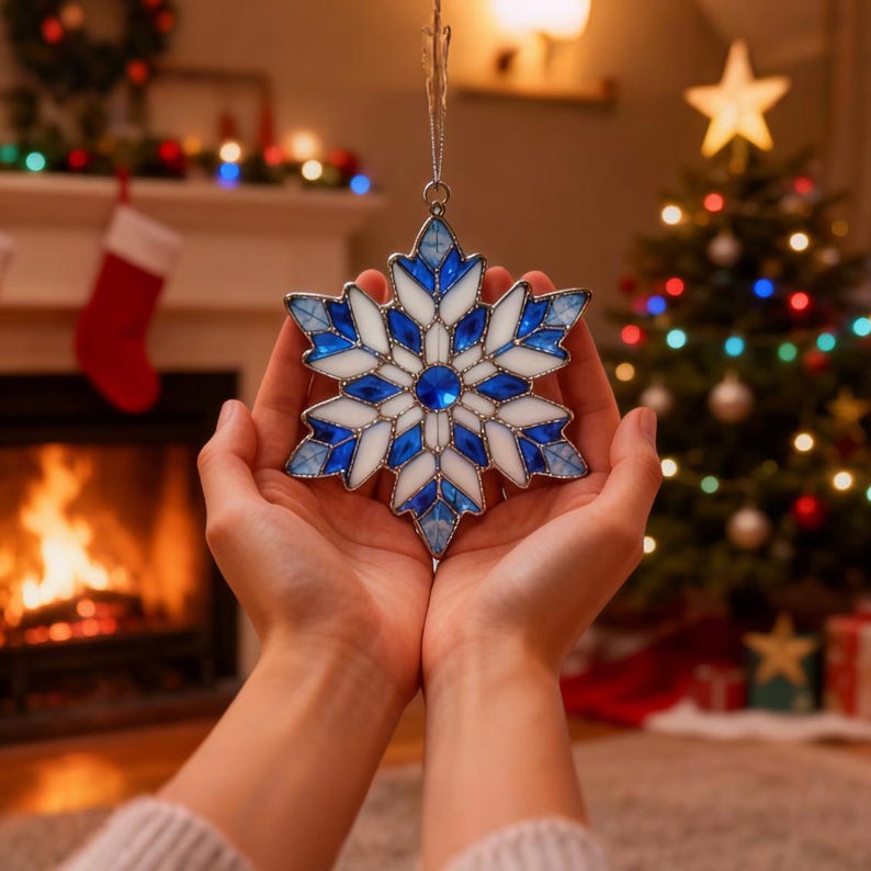 May include: A stained glass snowflake ornament with blue and white segments, held in cupped hands. The ornament has a silver hanging loop. The background includes a Christmas tree with lights and a fireplace with a fire burning.
