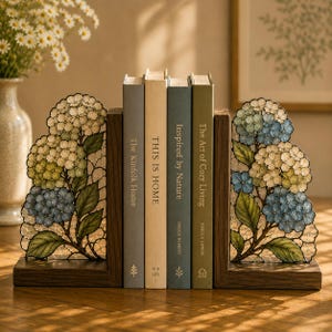 以下が含まれることがあります： 青、クリーム、緑の色合いでアジサイの花が描かれた、ステンドグラス風の装飾パネル付きL字型木製ブックエンド一対。ブックエンドの間には「The Kinfolk Home」、「This Is Home」、「Inspired by Nature」、「The Art of Cozy Living」というタイトルの本が4冊並んでいる。