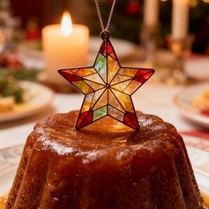 May include: A star-shaped ornament with stained glass design in red, yellow, green, and orange hues, suspended above a glazed cake. A lit candle and other festive decorations are in the background, suggesting a holiday theme.