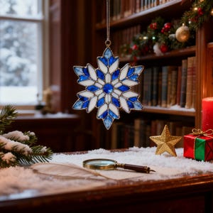 May include: A stained glass snowflake ornament with blue and white segments hangs in front of a bookshelf. A gold star, a magnifying glass, and a wrapped gift box sit on a snowy surface. A red candle and Christmas decorations are also visible.