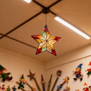 May include: A colorful, stained-glass star-shaped pendant light fixture. The star has a red border and is filled with various colors, including yellow, orange, and green. It hangs from a chain attached to a black ceiling fixture. The room is decorated with Christmas ornaments.