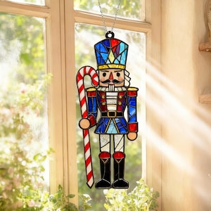 Puede incluir: Adorno navideño de vidriera de un soldado cascanueces. El adorno presenta un uniforme azul, rojo y dorado, un bigote negro y un bastón de caramelo. Cuelga de una cadena plateada frente a una ventana.
