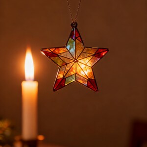 May include: A stained glass star ornament with a warm, glowing interior. The star is suspended by a gold chain and features red, orange, yellow, and green glass panels. A lit candle is in the background, casting a soft light.