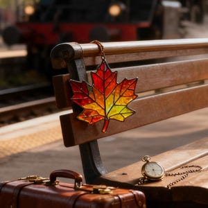 Op de afbeelding: Een gebrandschilderd esdoornbladornament in herfstkleuren rood, oranje en geel hangt aan een houten bank. Een vintage zakhorloge en een bruine koffer liggen op de bank. Op de achtergrond is een trein te zien.