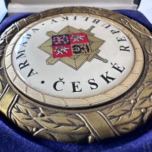 Op de afbeelding: Een ronde medaille in antieke stijl met de woorden "ARMADA REPUBLIKY ČESK&Eacute;" in zwarte letters. Het midden heeft een wit embleem met een gouden ster, zwaarden en een wapenschild. De medaille wordt gepresenteerd in een blauwe fluwelen doos.