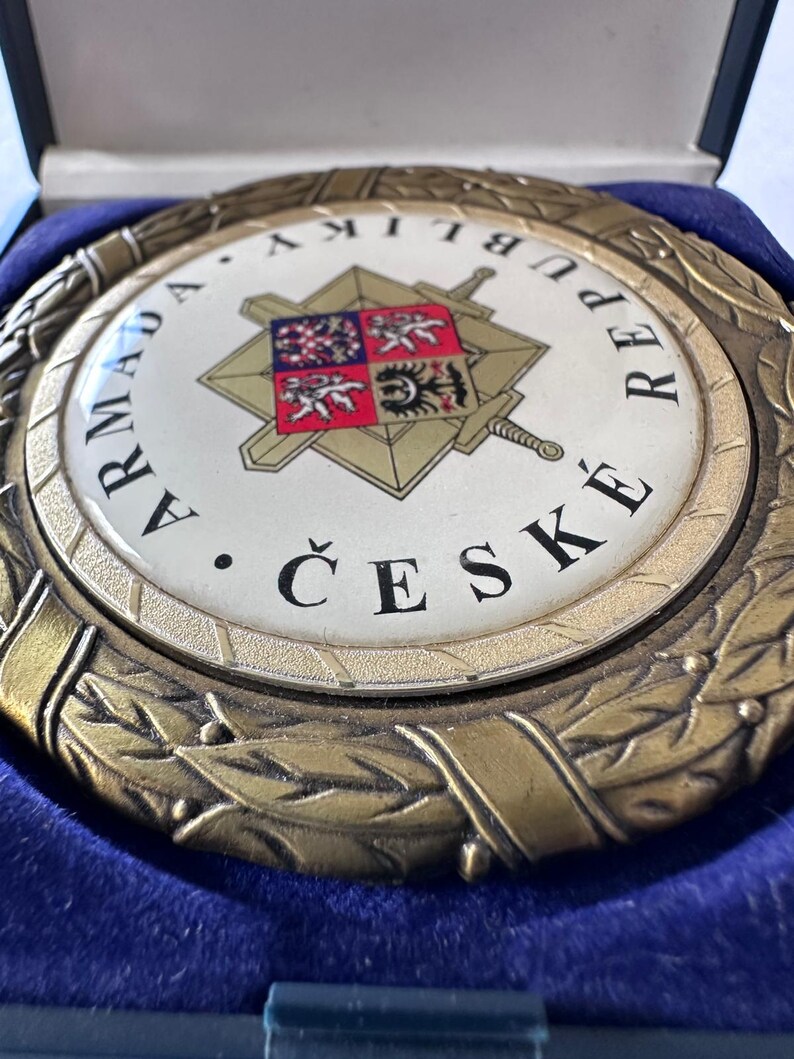 Op de afbeelding: Een ronde medaille in antieke stijl met de woorden "ARMADA REPUBLIKY ČESK&Eacute;" in zwarte letters. Het midden heeft een wit embleem met een gouden ster, zwaarden en een wapenschild. De medaille wordt gepresenteerd in een blauwe fluwelen doos.