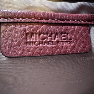 May include: Close-up of a burgundy leather item with the embossed text "MICHAEL MICHAEL KORS". The leather has a textured appearance, and the stitching is visible along the edges. The background is a light gray fabric.