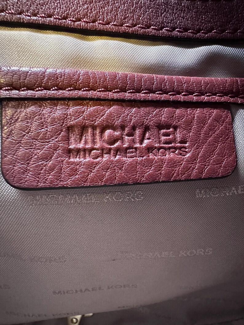 May include: Close-up of a burgundy leather item with the embossed text "MICHAEL MICHAEL KORS". The leather has a textured appearance, and the stitching is visible along the edges. The background is a light gray fabric.