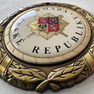 Op de afbeelding: Een cirkelvormig embleem in antieke stijl met de tekst "ČESK&Eacute; REPUBLIKY ARMADA" in zwarte letters. Het embleem heeft een centraal ontwerp met een wapenschild, gekruiste zwaarden en een gouden lauwerkrans.