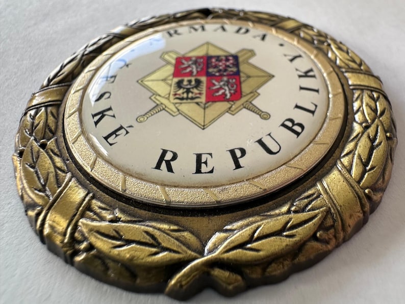 Op de afbeelding: Een cirkelvormig embleem in antieke stijl met de tekst "ČESK&Eacute; REPUBLIKY ARMADA" in zwarte letters. Het embleem heeft een centraal ontwerp met een wapenschild, gekruiste zwaarden en een gouden lauwerkrans.
