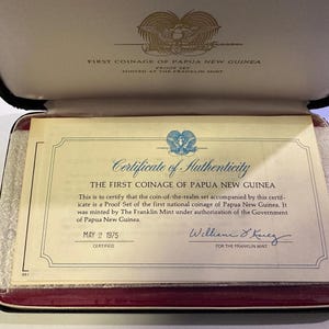Pode incluir: Certificado de autenticidade da primeira cunhagem da Papua Nova Guin&eacute;, datado de 2 de maio de 1975. O documento est&aacute; dentro de um estojo preto com interior vermelho. O texto diz "Primeira cunhagem da Papua Nova Guin&eacute;".