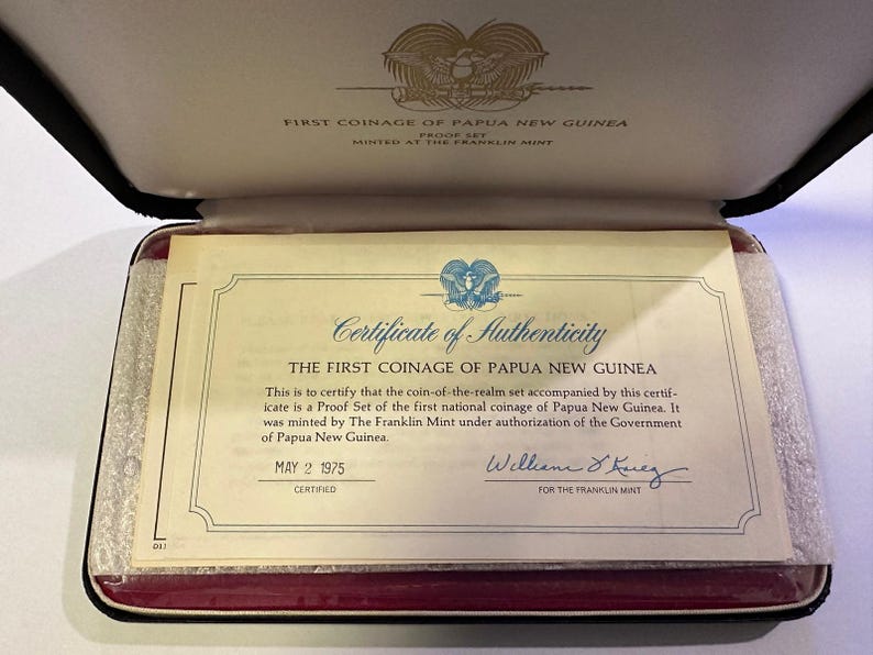 Pode incluir: Certificado de autenticidade da primeira cunhagem da Papua Nova Guin&eacute;, datado de 2 de maio de 1975. O documento est&aacute; dentro de um estojo preto com interior vermelho. O texto diz "Primeira cunhagem da Papua Nova Guin&eacute;".