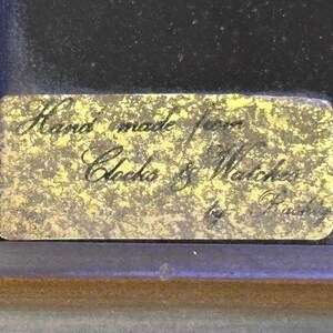 May include: A rectangular brass plaque with the inscription "Hand made from Clocks & Watches by Rusty" in elegant script. The plaque is set against a dark background within a wooden frame.