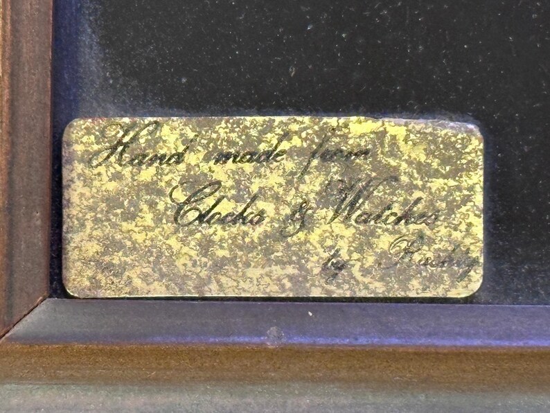 May include: A rectangular brass plaque with the inscription "Hand made from Clocks & Watches by Rusty" in elegant script. The plaque is set against a dark background within a wooden frame.