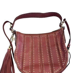 May include: A burgundy leather shoulder bag with a curved shape. The bag features vertical rows of gold-colored eyelets and a matching tassel. The strap and trim are also burgundy, with gold hardware and a chain detail.