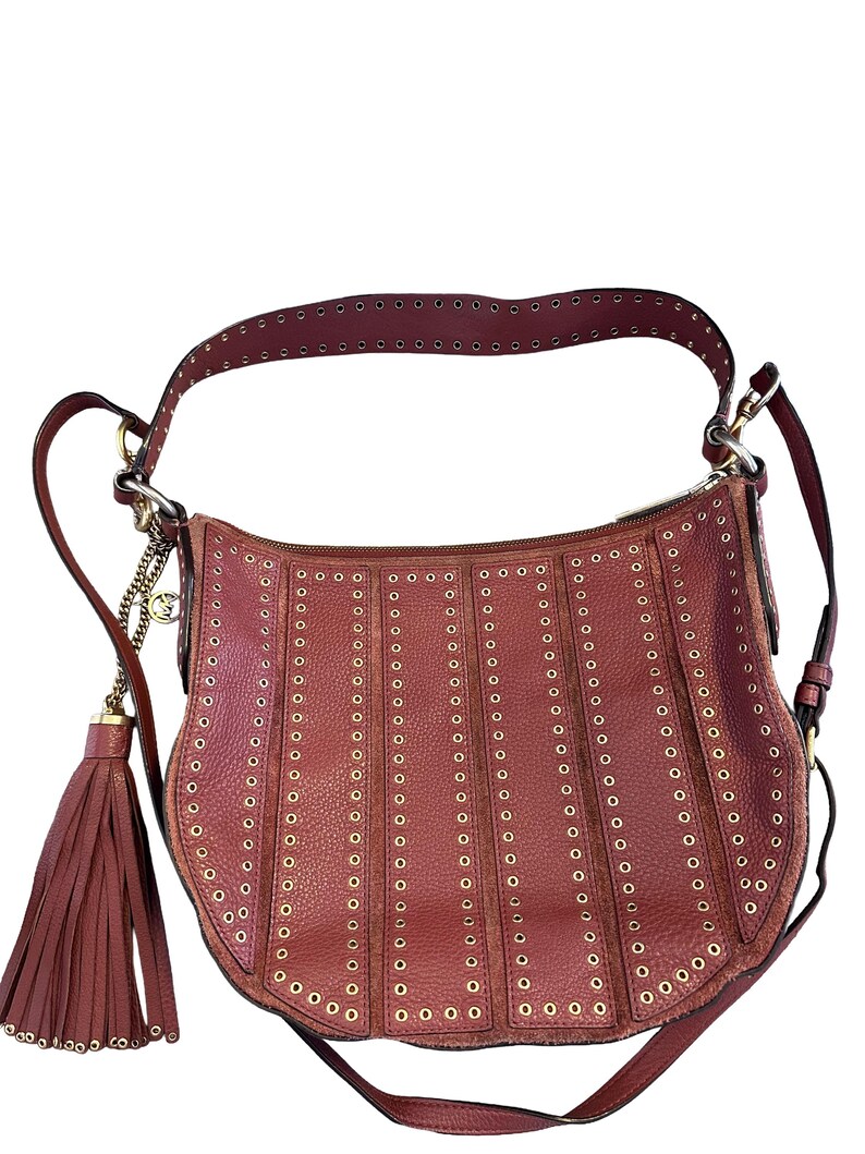 May include: A burgundy leather shoulder bag with a curved shape. The bag features vertical rows of gold-colored eyelets and a matching tassel. The strap and trim are also burgundy, with gold hardware and a chain detail.