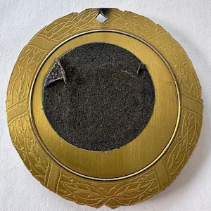 Op de afbeelding: Een ronde, antiek goudkleurige lijst met een decoratief bladerpatroon langs de rand. Het midden heeft een donkergrijze viltrug met een cirkelvormige opening. Er zit een klein, ruitvormig gaatje aan de bovenkant.