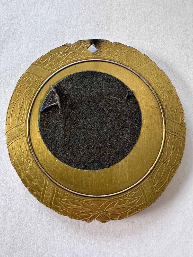 Op de afbeelding: Een ronde, antiek goudkleurige lijst met een decoratief bladerpatroon langs de rand. Het midden heeft een donkergrijze viltrug met een cirkelvormige opening. Er zit een klein, ruitvormig gaatje aan de bovenkant.
