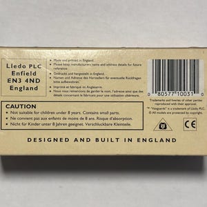 以下が含まれることがあります： 「Lledo PLC Enfield EN3 4ND England」と「Designed and Built in England」などのテキストが記載された、ベージュ色の段ボール箱。 バーコードと小さな部品に関する警告があります。 この箱は玩具用です。