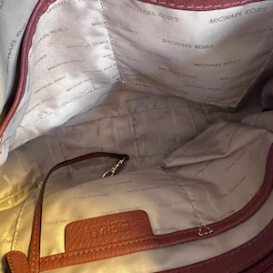 May include: Interior view of a burgundy Michael Kors handbag. The lining is a light beige color with the Michael Kors logo printed repeatedly. A small leather tag with the Michael Kors name is visible. A strap and a gold-colored clasp are also visible.