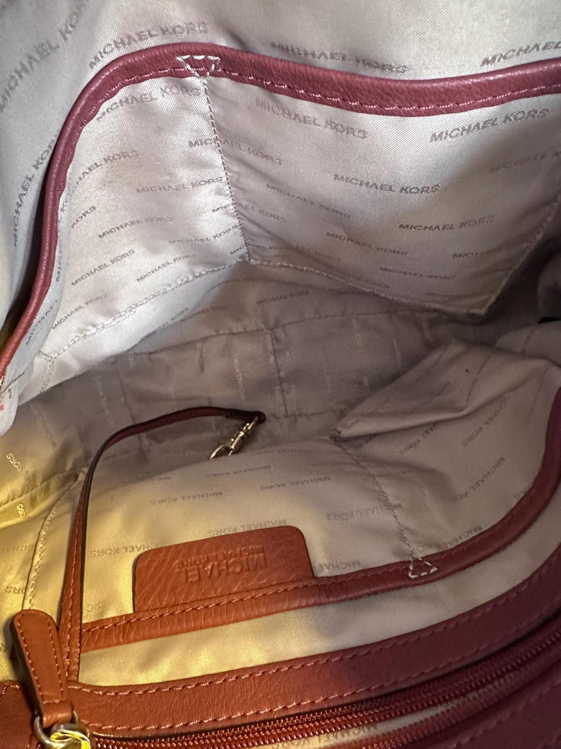 May include: Interior view of a burgundy Michael Kors handbag. The lining is a light beige color with the Michael Kors logo printed repeatedly. A small leather tag with the Michael Kors name is visible. A strap and a gold-colored clasp are also visible.