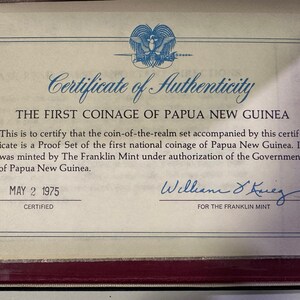 Pode incluir: Certificado de autenticidade da primeira cunhagem da Papua Nova Guin&eacute;, datado de 2 de maio de 1975. O documento certifica um Proof Set cunhado pela The Franklin Mint. Apresenta um emblema azul e uma assinatura.