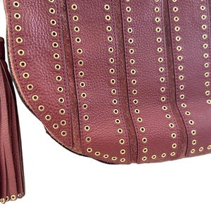 May include: Close-up of a burgundy leather handbag with gold-tone grommets and a matching tassel. The bag features vertical rows of grommets and a gold chain strap.