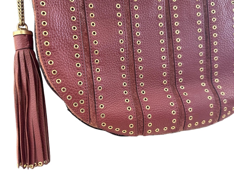 May include: Close-up of a burgundy leather handbag with gold-tone grommets and a matching tassel. The bag features vertical rows of grommets and a gold chain strap.
