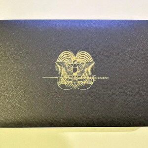 Pode incluir: Uma caixa retangular preta com um emblema dourado com um p&aacute;ssaro e uma espada. A caixa tem uma superf&iacute;cie texturizada e cantos arredondados. O emblema est&aacute; centrado na tampa.