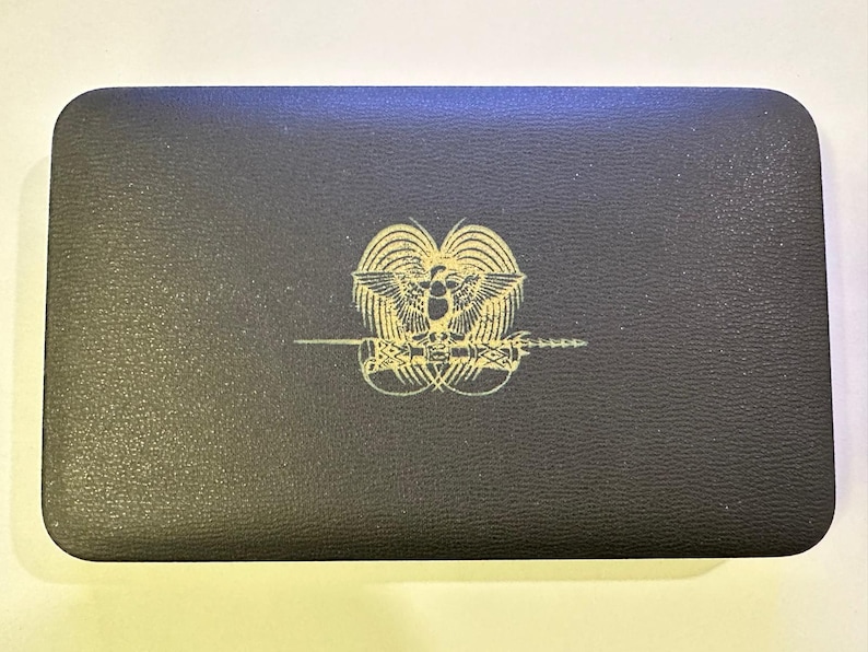 Pode incluir: Uma caixa retangular preta com um emblema dourado com um p&aacute;ssaro e uma espada. A caixa tem uma superf&iacute;cie texturizada e cantos arredondados. O emblema est&aacute; centrado na tampa.