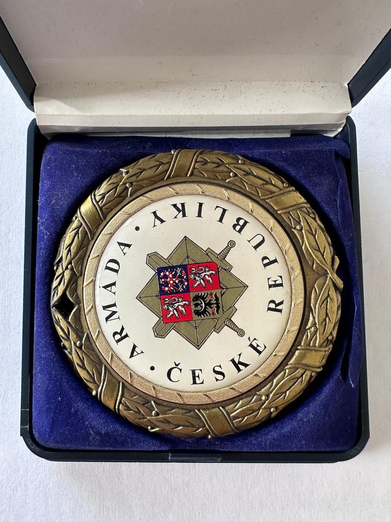 Op de afbeelding: Ronde, bronskleurige medaille met een wit midden, met een wapen en Tsjechische tekst. De medaille is omgeven door een laurierkrans en wordt gepresenteerd in een zwarte doos met een blauwe fluwelen voering.